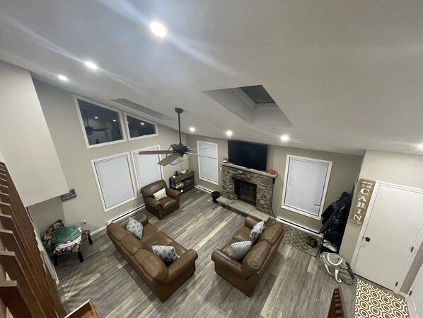 A spacious living room with a fireplace, two sofas, a ceiling fan, and a TV mounted above the fireplace.