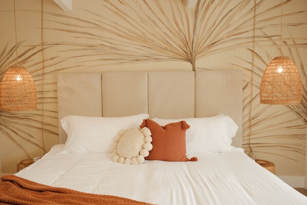 A neatly made bed with white linens and a brown throw blanket, a round pillow with a pom-pom border, and a brown pillow. There are two woven light fixtures on either side of the bed. - The Maya (Palm Springs)