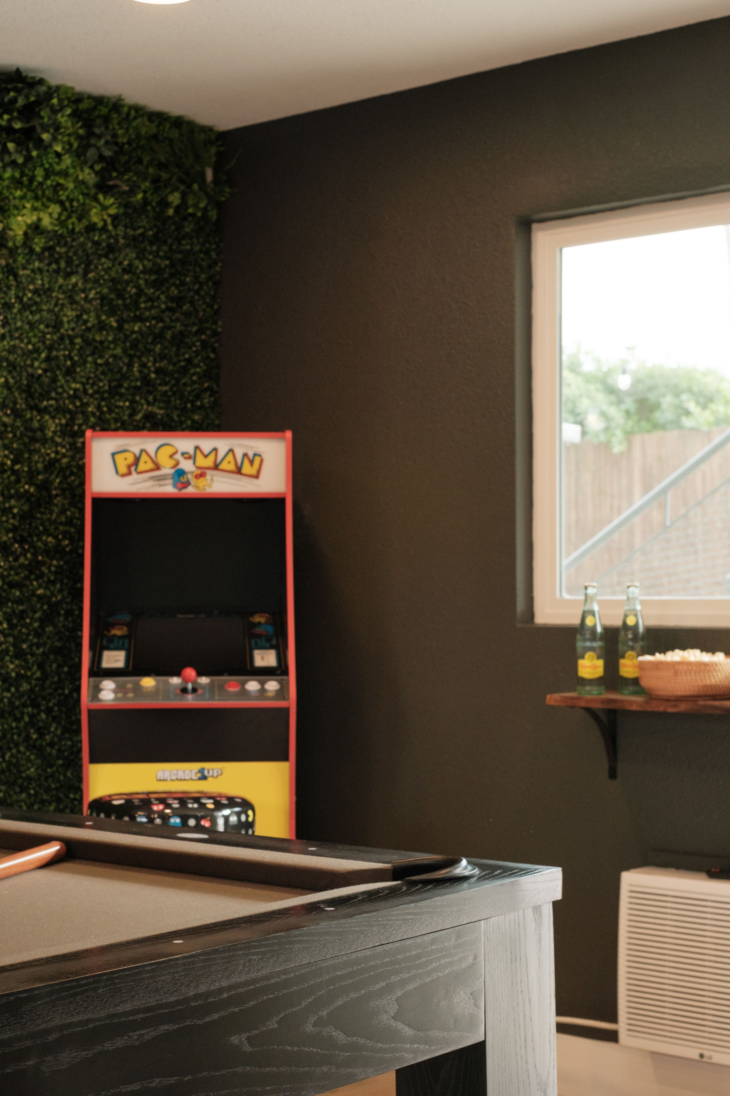 Ein Billardtisch, ein Pac-Man-Spielautomat, ein Fenster mit Blick auf einen Holenzaun und ein Regal mit Flaschen.