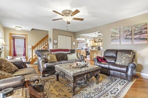 A spacious living room with leather sofas, a large ornate coffee table, and a patterned rug. A ceiling fan and a staircase are visible in the background.