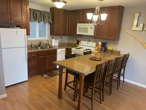 A kitchen with wooden cabinets, a white refrigerator, a microwave, a stove, and a dining area with a granite countertop and wooden chairs.