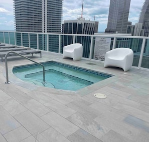 A rooftop pool with a view of city buildings, two white lounge chairs, and a sign about spa rules.
