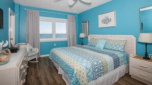 Un dormitorio con cama, cómoda, tocador, ventilador de techo, ventana con cortinas e imagen enmarcada en la pared.