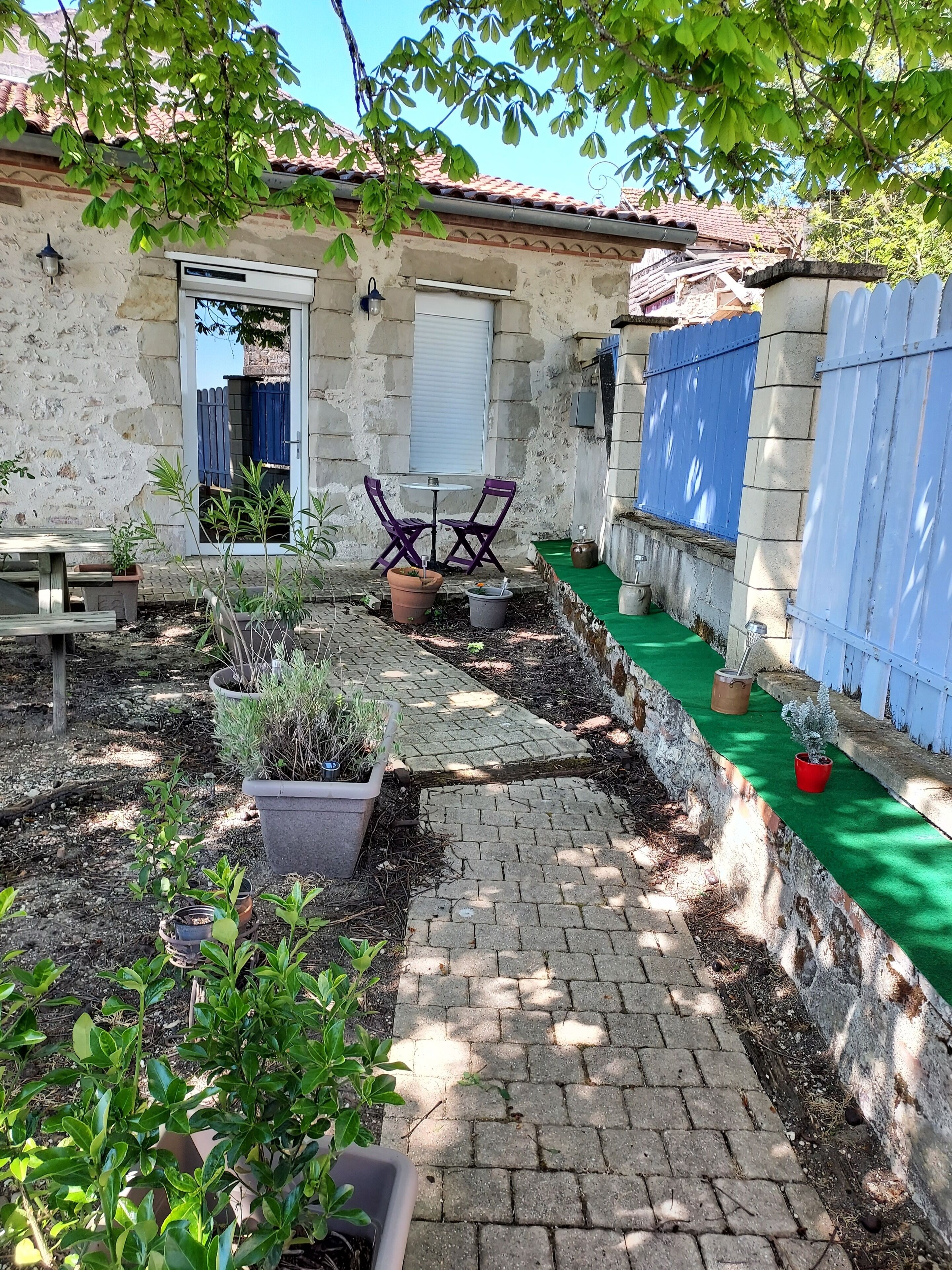 A courtyard with a paved pathway, potted plants, a table with chairs, and a blue fence.