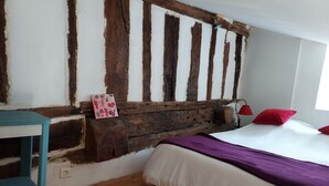 A bedroom with exposed wooden beams, a bed with white and purple bedding, a small table, and a red lamp.
