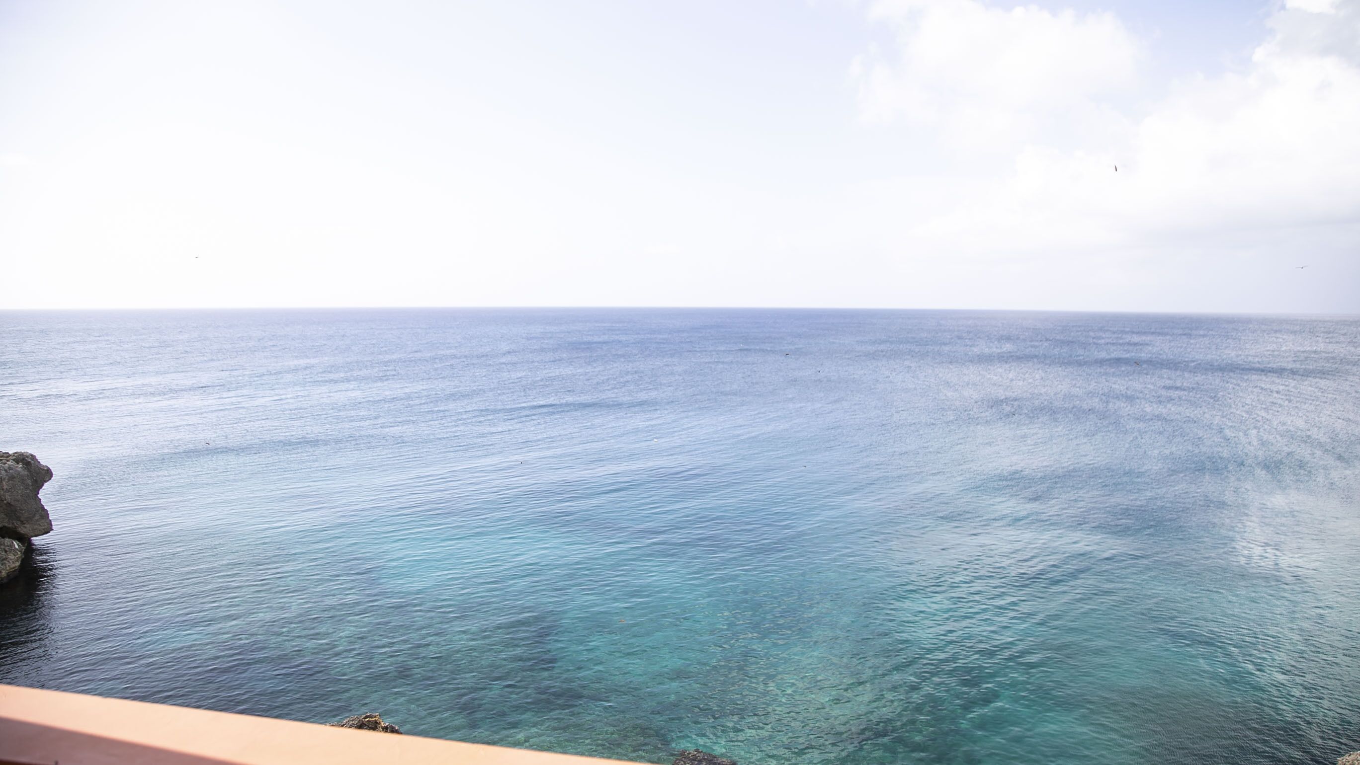 A clear view of the sea from a balcony with a rock formation on the left.
