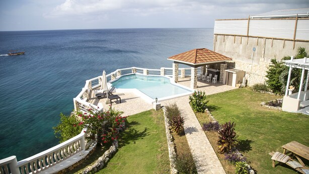 Une piscine entourée d’une terrasse en pierre, elle-même bordée d’un muret en pierre et d’une clôture blanche. On y trouve également un espace patio avec des meubles d’extérieur et une pergola.