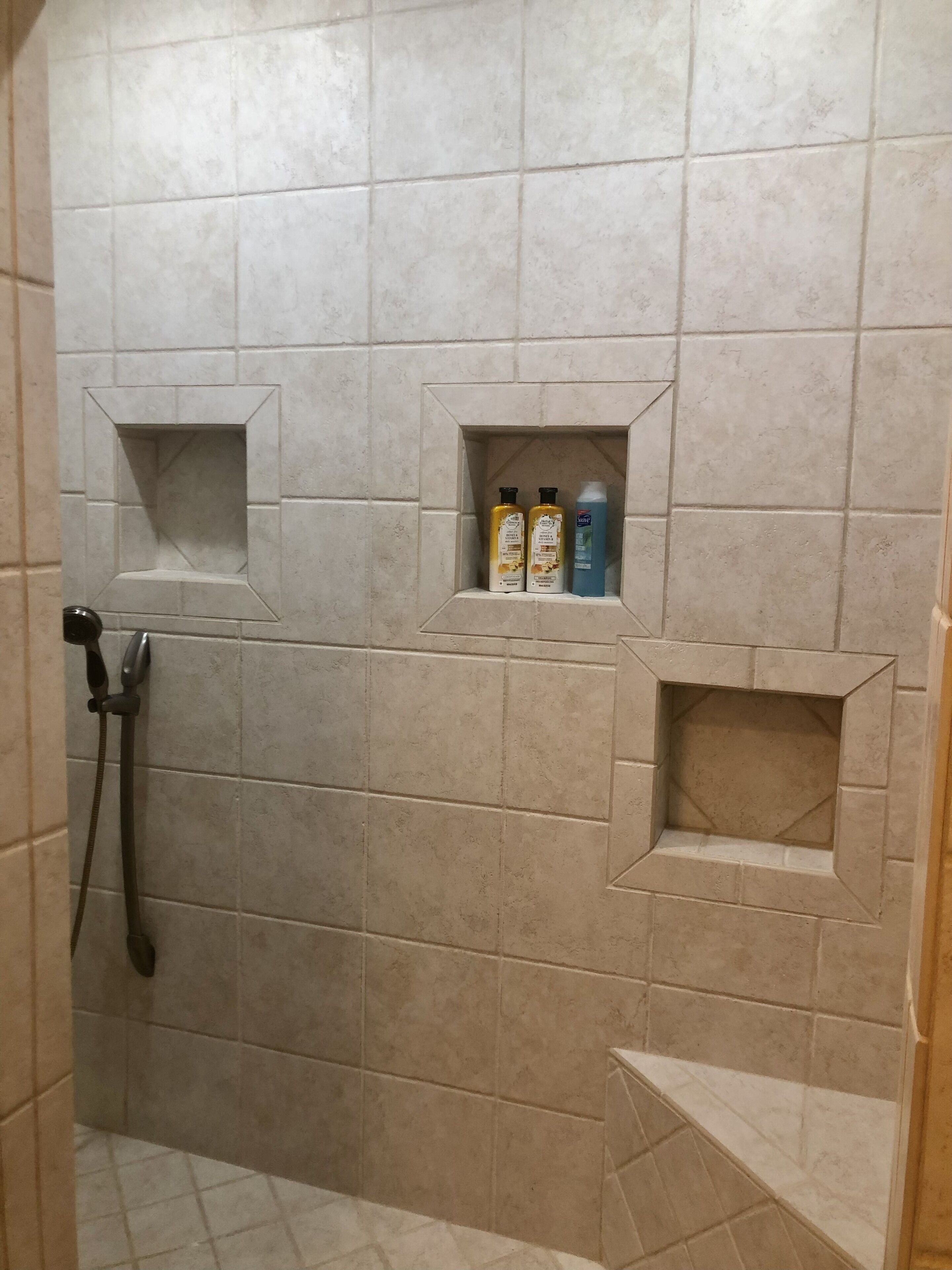 Un espace douche avec des murs carrelés, une douchette à main et deux étagères contenant des articles de toilette.