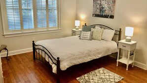 Un dormitorio ordenado con cama, mesitas de noche, un velador y una pintura en la pared.