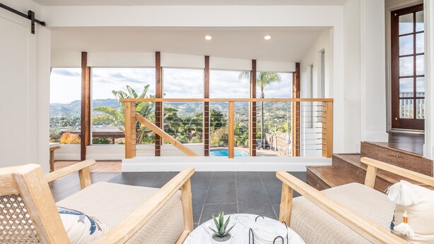 A spacious balcony with a view of mountains and a pool, furnished with wooden chairs and a marble table.