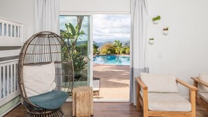 A room with a wicker chair, a wooden bench, and a view of a swimming pool and mountains.