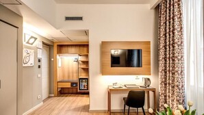 Une chambre d’hôtel avec un bureau en bois, un téléviseur à écran plat fixé au mur, une petite kitchenette et une fenêtre agrémentée de rideaux à motifs.