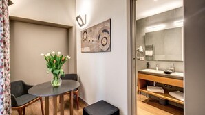 Une chambre d’hôtel moderne avec une table de salle à manger ronde, un vase de fleurs, un grand tableau abstrait et une salle de bain dotée d’un meuble-lavabo en bois.