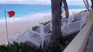 A staircase leading to a beach with a red flag, white railings, and palm trees. - Sunrise Beach Villas #2.. Oceanfront Villa (New Providence)