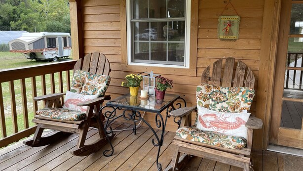 A wooden porch with two rocking chairs and a table, a window, and a camper parked outside.