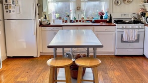 A kitchen with a white refrigerator, stove, and sink, wooden stools, and a hanging light fixture.