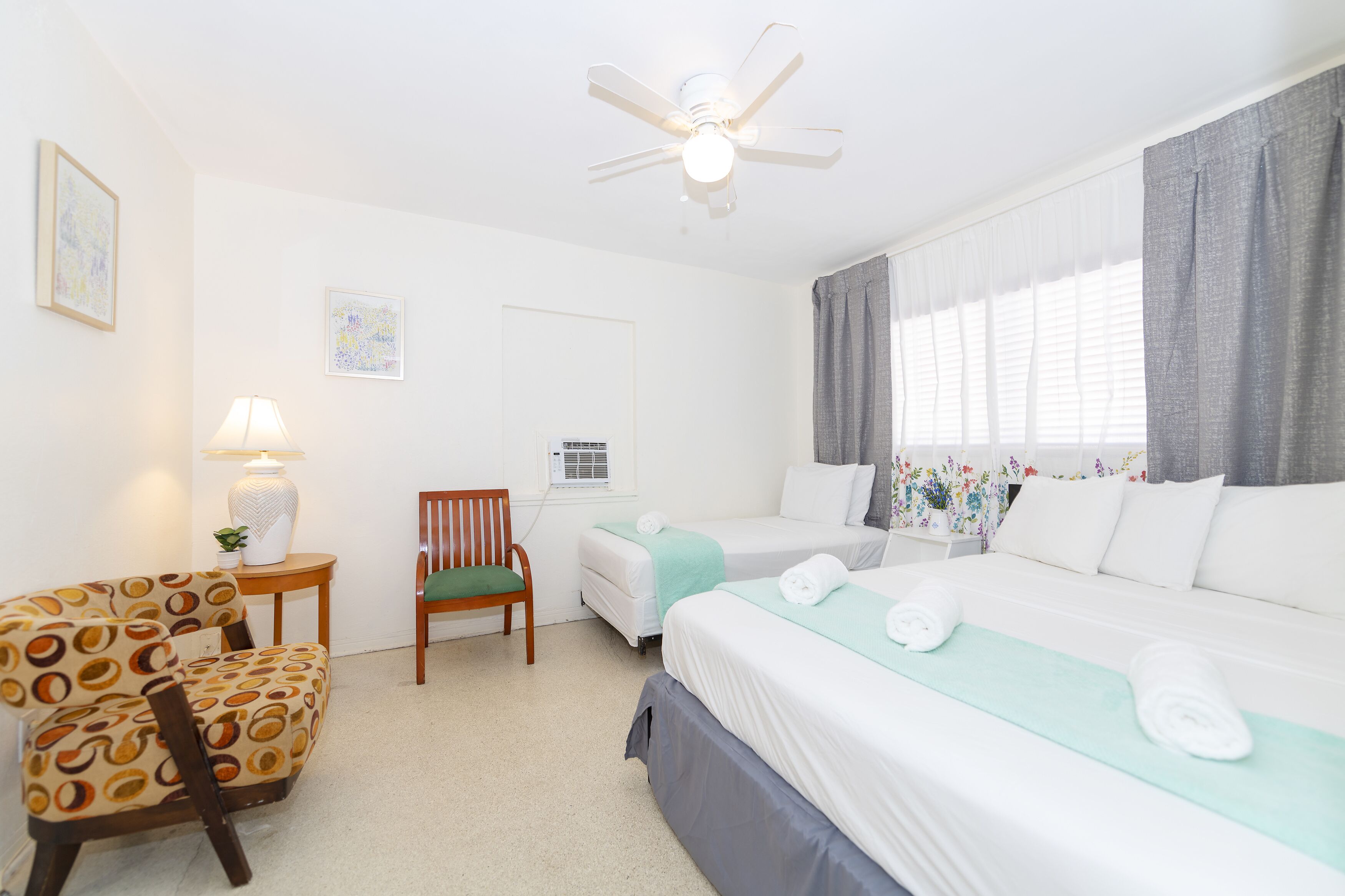 Habitación de hotel con dos camas, una silla, una lámpara y un ventilador de techo.
