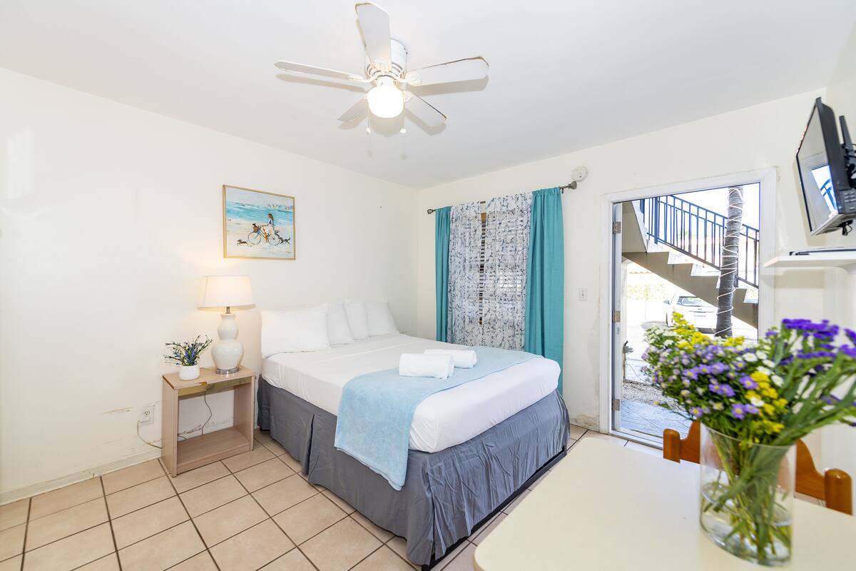 Ein Hotelzimmer mit Bett, Nachttisch, Lampe, Deckenventilator, einem Fenster mit Vorhängen, einer Vase mit Blumen und einem Fernseher an der Wand.