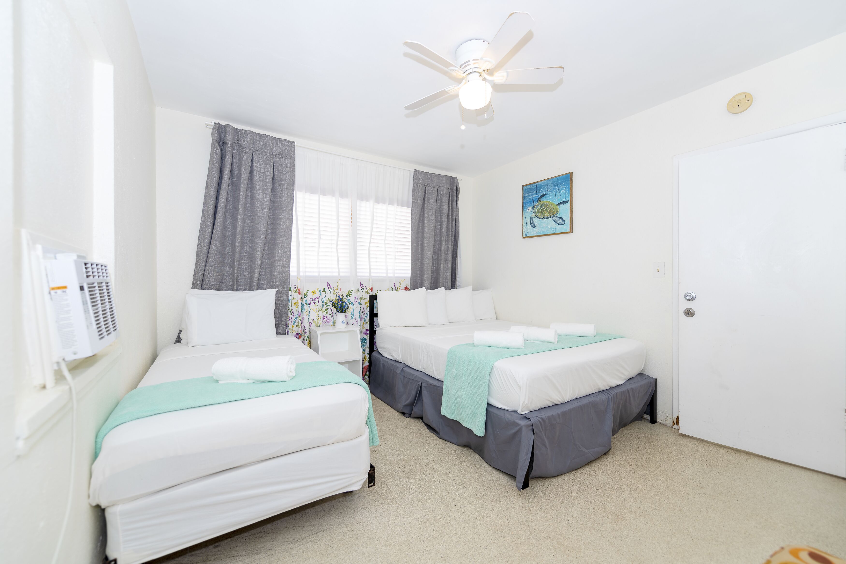 A hotel room with two beds, a ceiling fan, a window with curtains, a painting, and a white door.