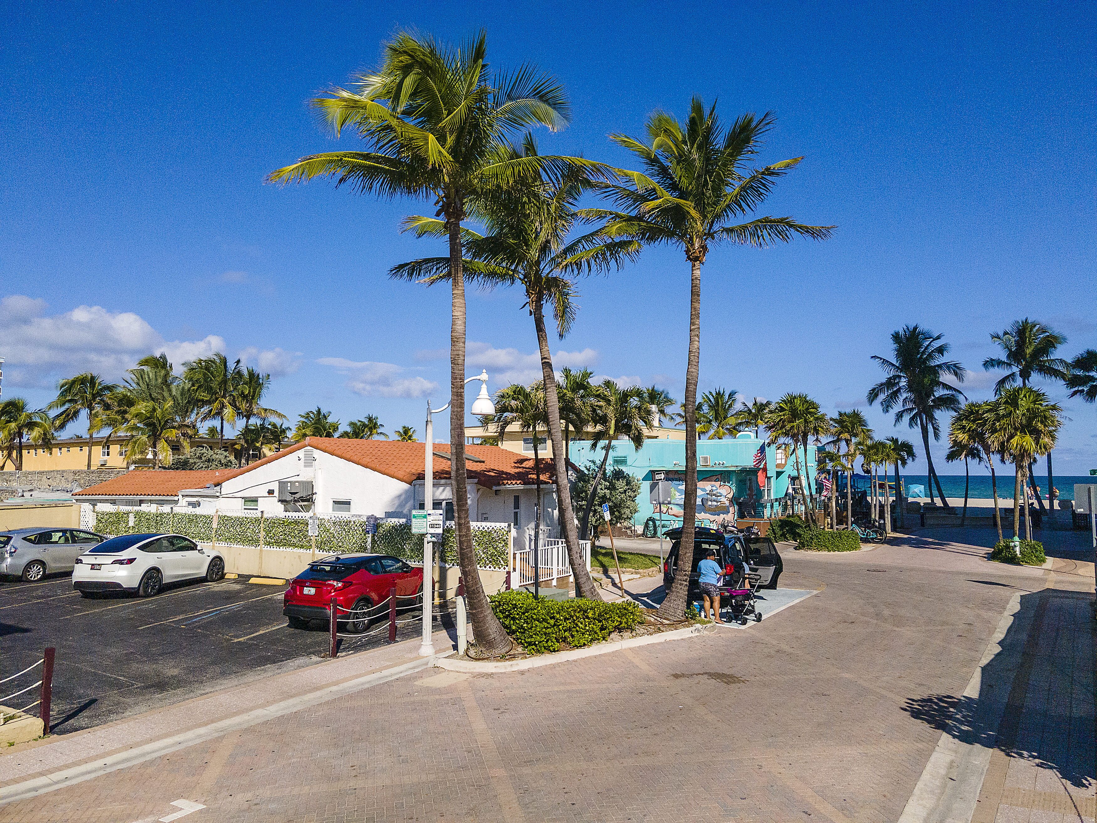 A street with parked cars, palm trees, and a building with a red roof.