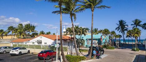 A street with parked cars, palm trees, and a building with a red roof.