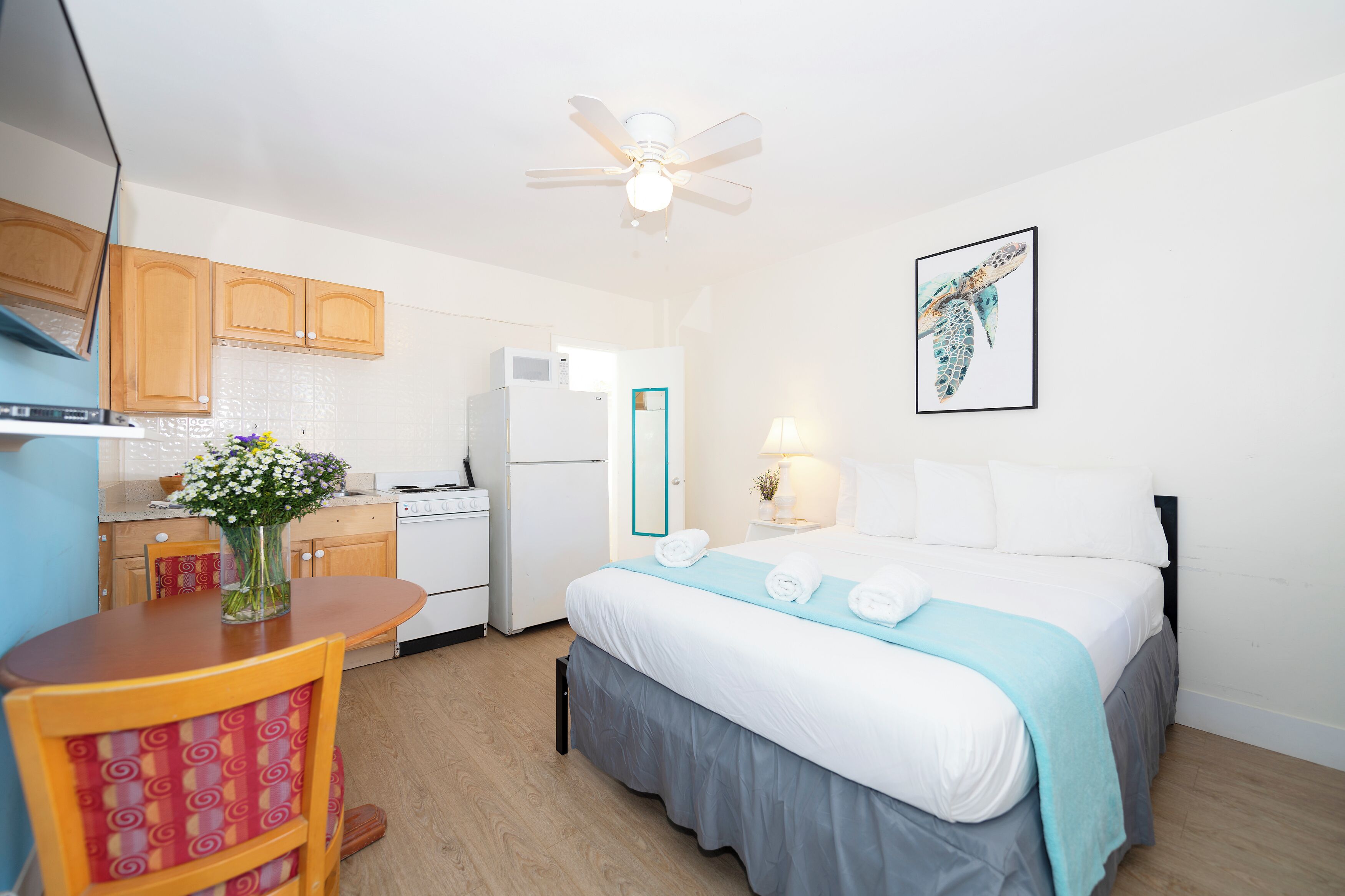 A compact hotel room with a bed, a small kitchenette, a dining area, and a ceiling fan.