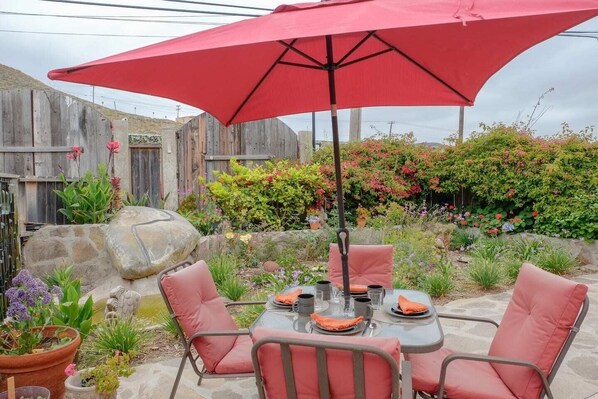 Une terrasse extérieure avec une table dressée pour deux personnes, entourée de chaises, et protégée par un grand parasol rouge.