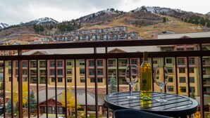 Ein Balkon mit Tisch, einer Weinflasche und zwei Weingläsern. Im Hintergrund ein mehrstöckiges Gebäude und eine gebirgige Landschaft.