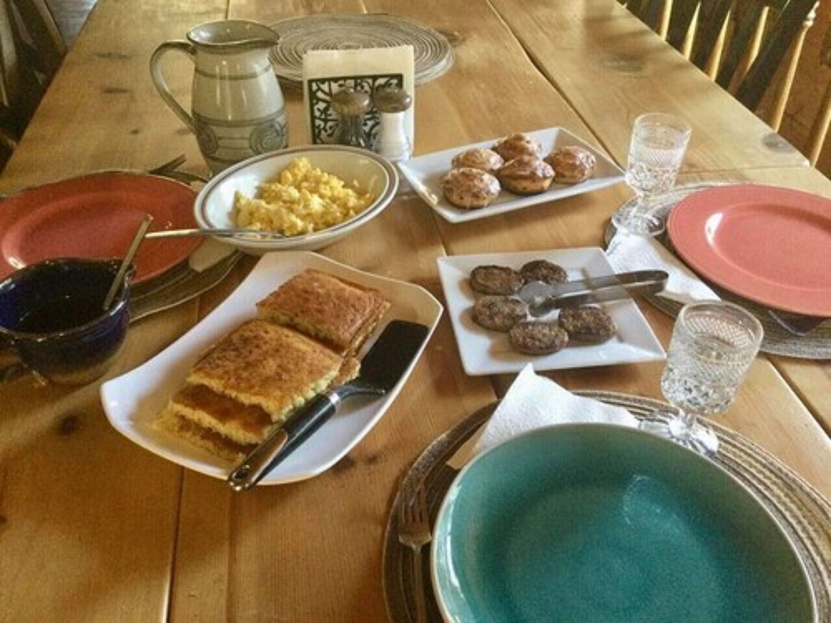 A wooden dining table with plates of food, including scrambled eggs, meatballs, and a slice of cake.
