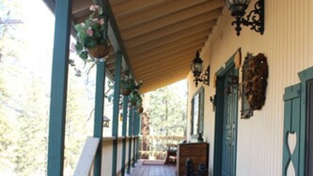 A wooden porch with steps leading to a door, a hanging flower basket, and a wall-mounted lantern. - The Old Bear BnB (Pine Mountain Club)