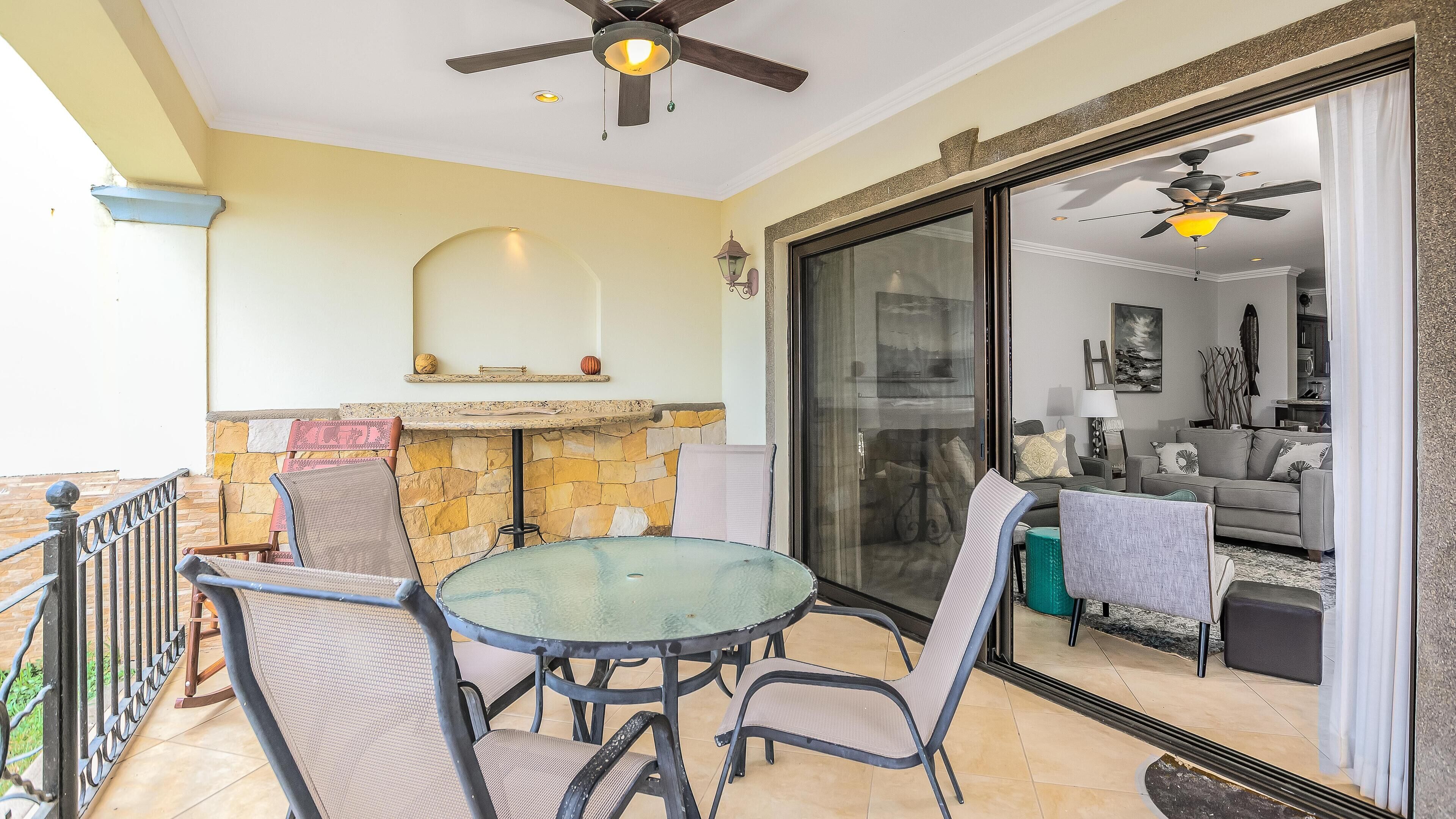 An outdoor patio with a round table and chairs, a stone fireplace, and a view into the living area through a glass door.