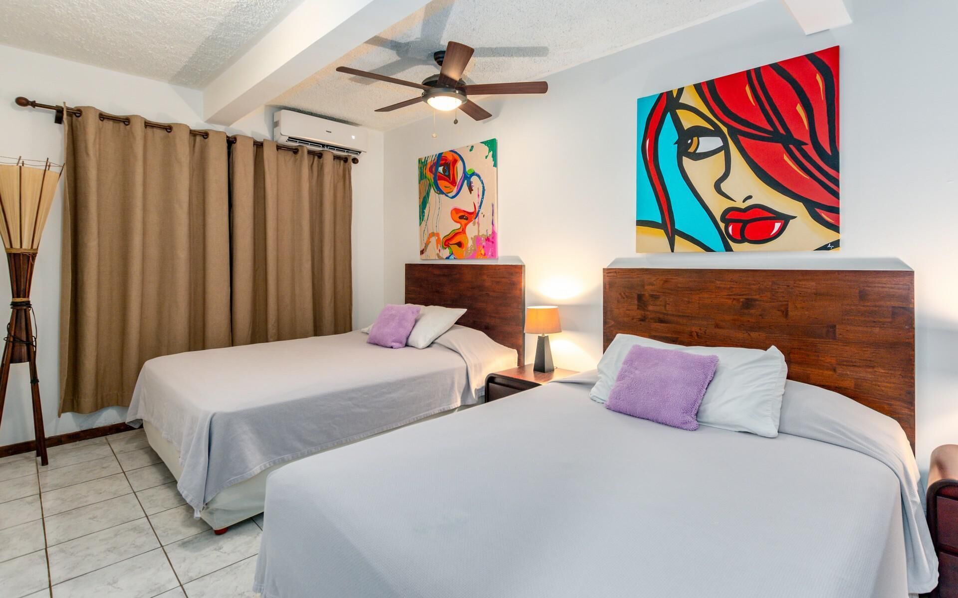 Habitación de hotel con dos camas, ventilador de techo, un cuadro en la pared y una lámpara en la mesita de noche.