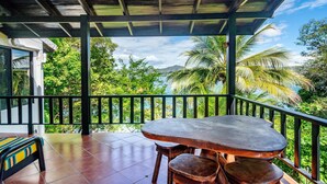 Un balcón techado con una mesa y sillas, con vista a un paisaje exuberante con palmeras y el mar.