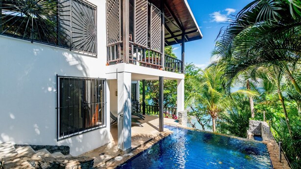 Una casa moderna de dos pisos con piscina, rodeada de abundante vegetación y palmeras.