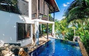 A modern two-story house with a swimming pool, surrounded by lush greenery and palm trees.