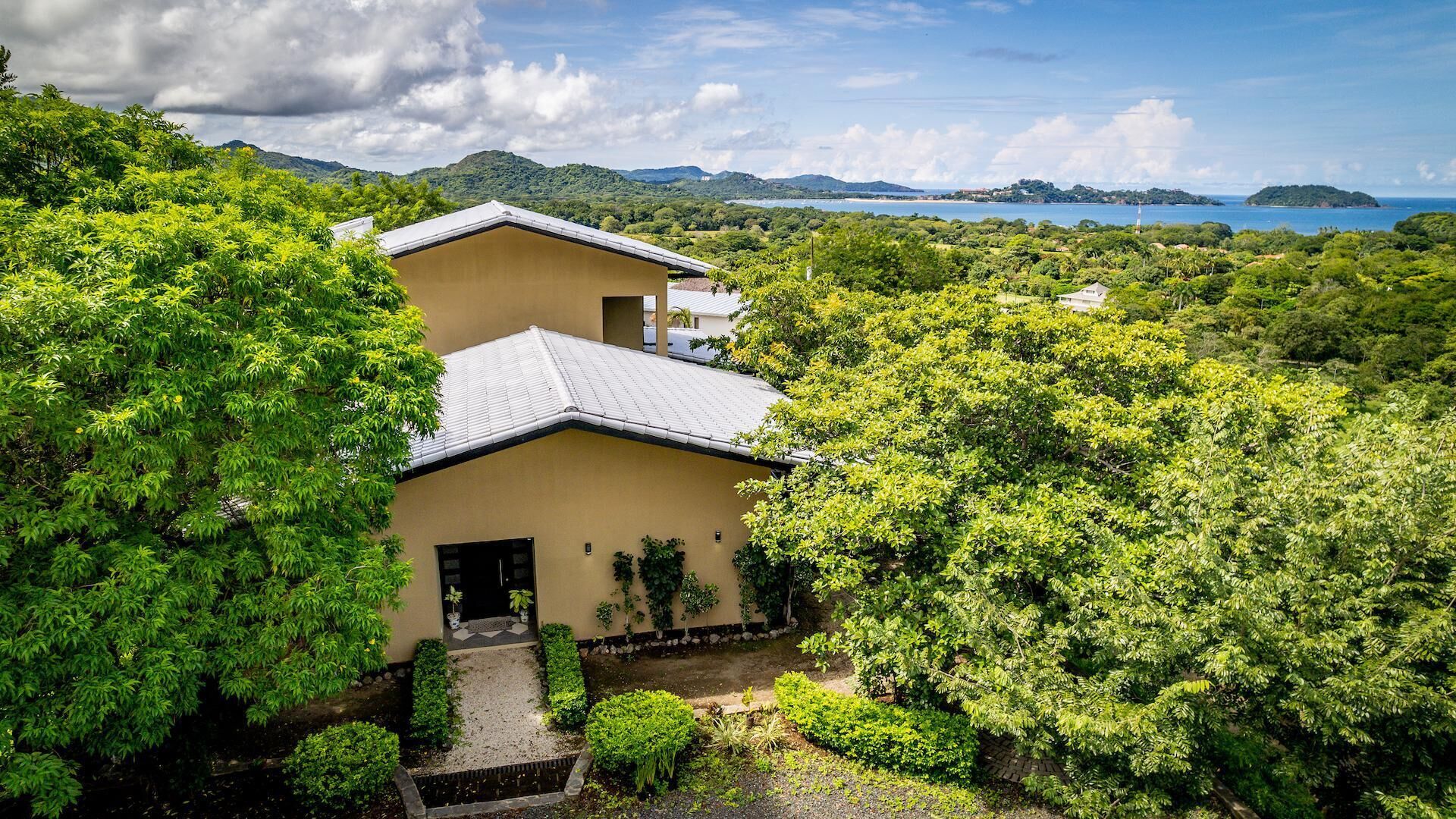 A two-story building surrounded by lush greenery, with a view of a coastline and distant islands.