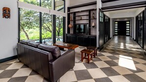 A modern living room with a leather sofa, a coffee table, and a TV. There is a large window with a view of greenery outside. - Nostalgic Retro Home on Luxurious Lot with a Private Infinity Pool and Ocean Views (Tempate)