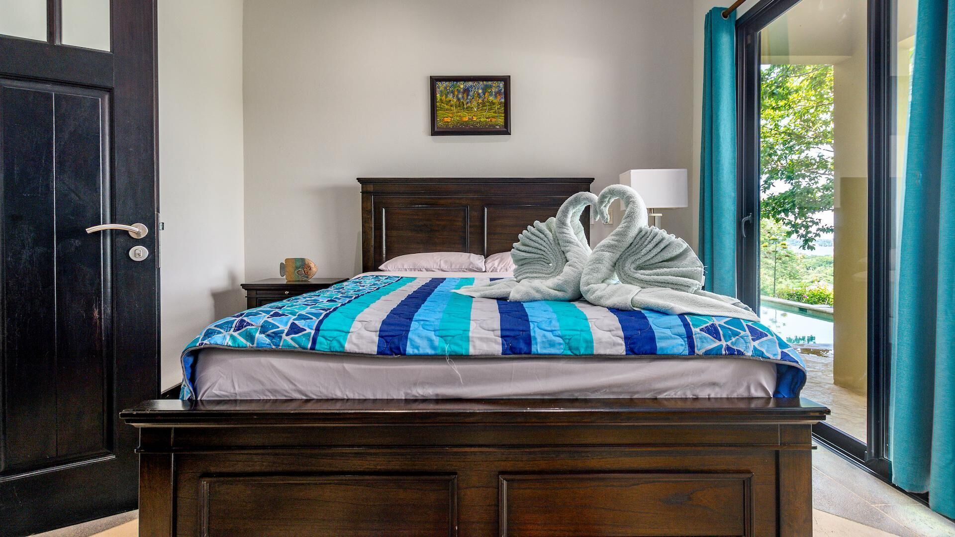 A bed with a blue and white quilt, a wooden headboard, a framed picture on the wall, and a view of greenery through a window.