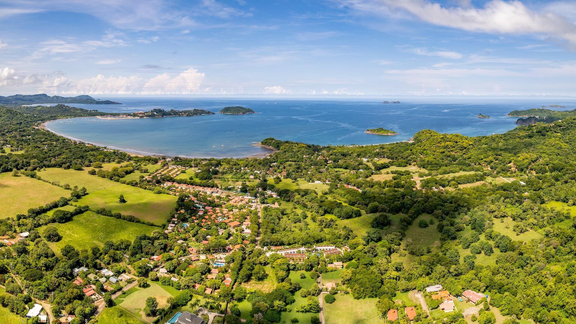 An aerial view of a coastal town with a mix of residential and commercial buildings, surrounded by lush greenery and a large body of water.