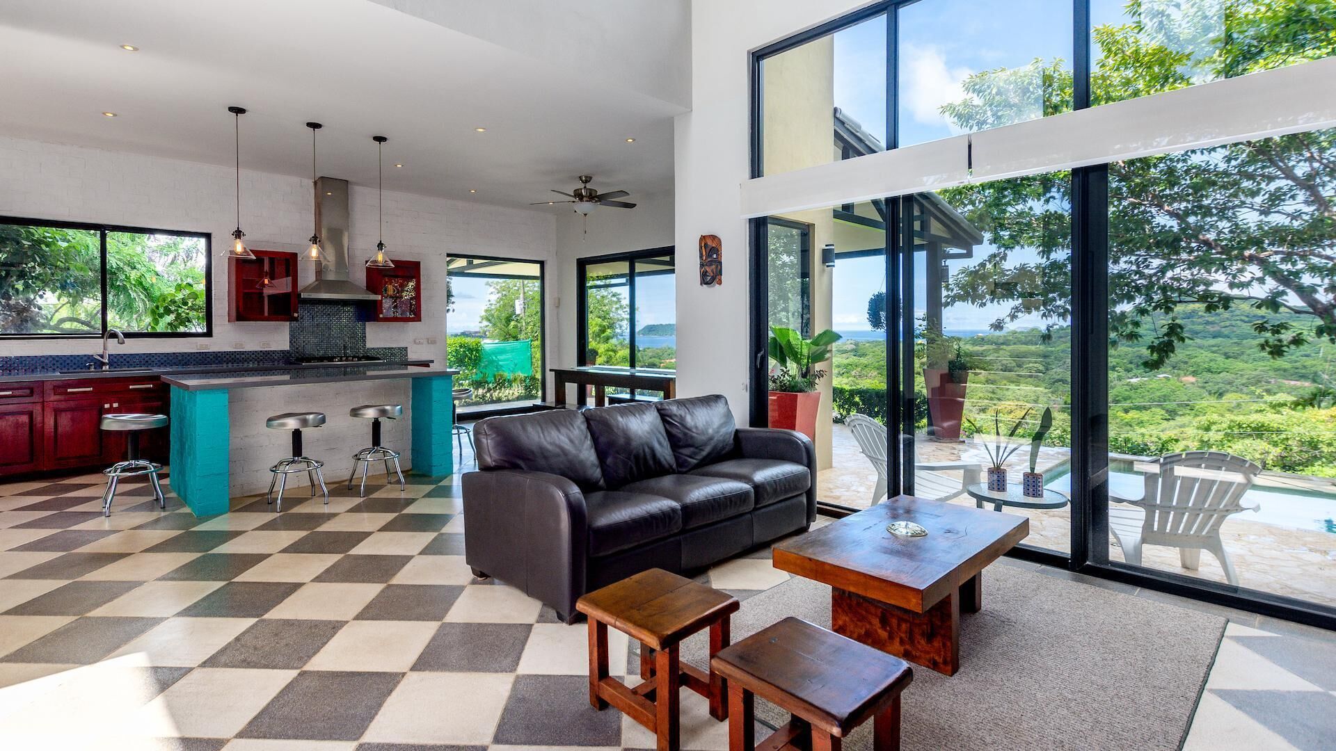 A modern kitchen with a large island, bar stools, and a black leather sofa. There is a dining area with a wooden table and chairs, and large windows offering a view of the outdoors.