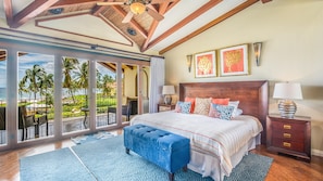 A spacious bedroom with a large bed, a wooden headboard, and a view of palm trees and the ocean through floor-to-ceiling windows.