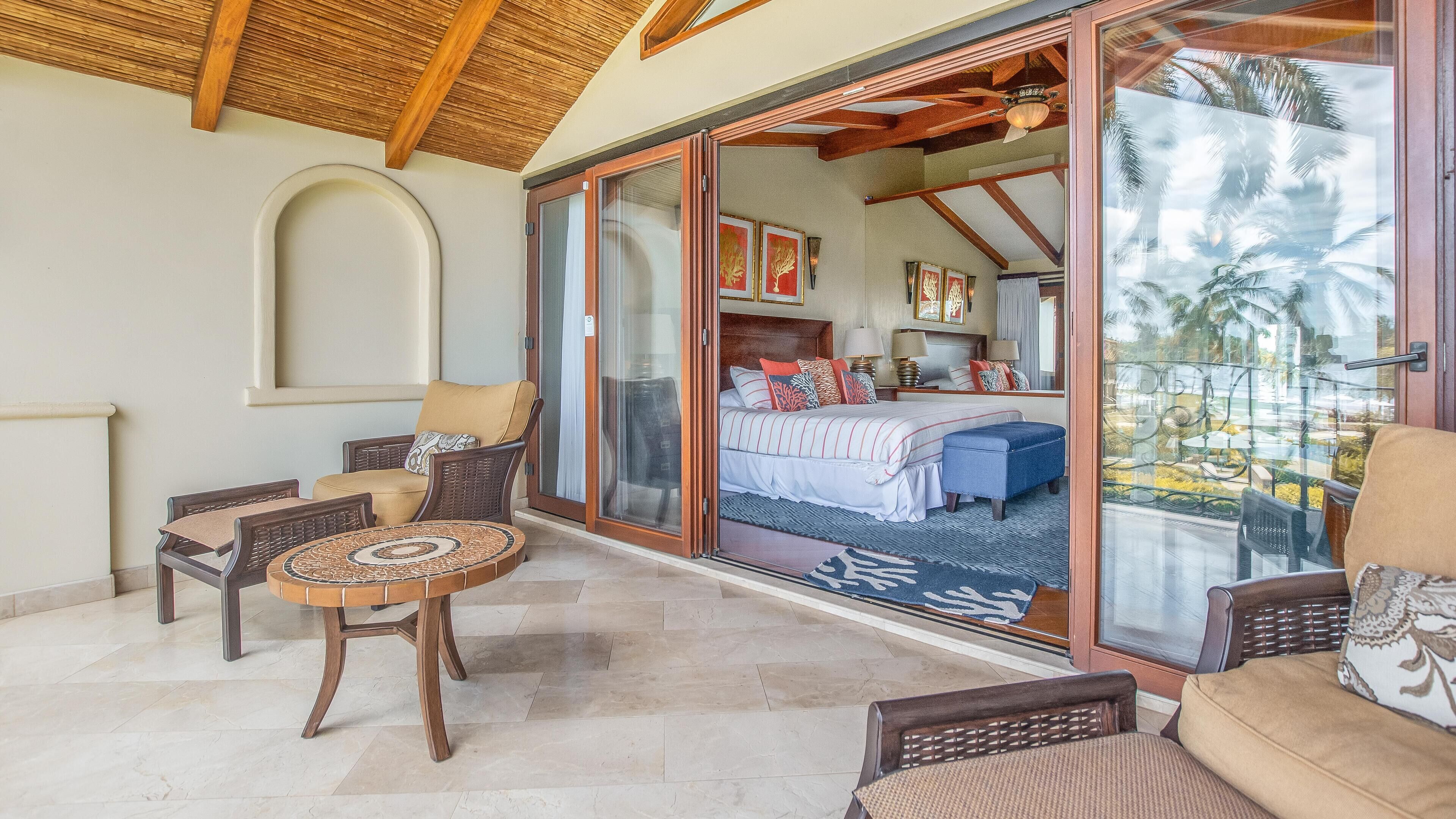 Ein Zimmer mit einem Bett, einem Sessel, einem Tisch und einer Glasschiebetür, die auf einen Balkon mit Blick auf Palmen führt.