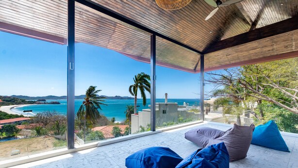A modern room with large glass windows overlooking a coastal view, featuring a wooden ceiling and minimalist furniture. - Luxury Ocean-View Flamingo Home with Pool, Apartment and Party Deck (Cabo Velas)