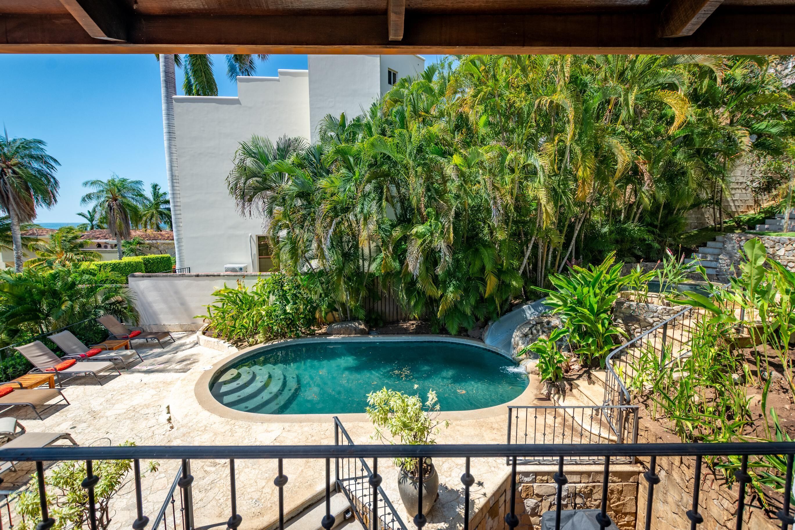 A pool area with lounge chairs, surrounded by lush greenery and a white building.