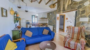 A cozy living room with a blue sofa, a striped armchair, and a small coffee table. A kitchen is visible in the background with a microwave and a window. - El Garrotal - El Pan El Bosque by Ruralidays (Cádiz)