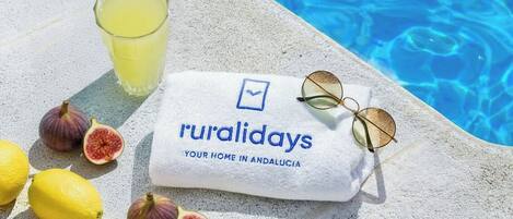 A towel with 'ruralidays' branding, lemons, figs, and a glass of yellow liquid by a pool.