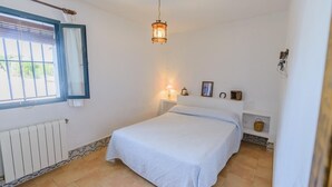 A bedroom with a bed, a window with curtains, a hanging light fixture, a radiator, and a wall-mounted shelf with items. - El Garrotal - El Pan El Bosque by Ruralidays (Cádiz)