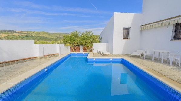 A rectangular swimming pool with blue tiles, surrounded by white walls and a small patio area with white chairs.