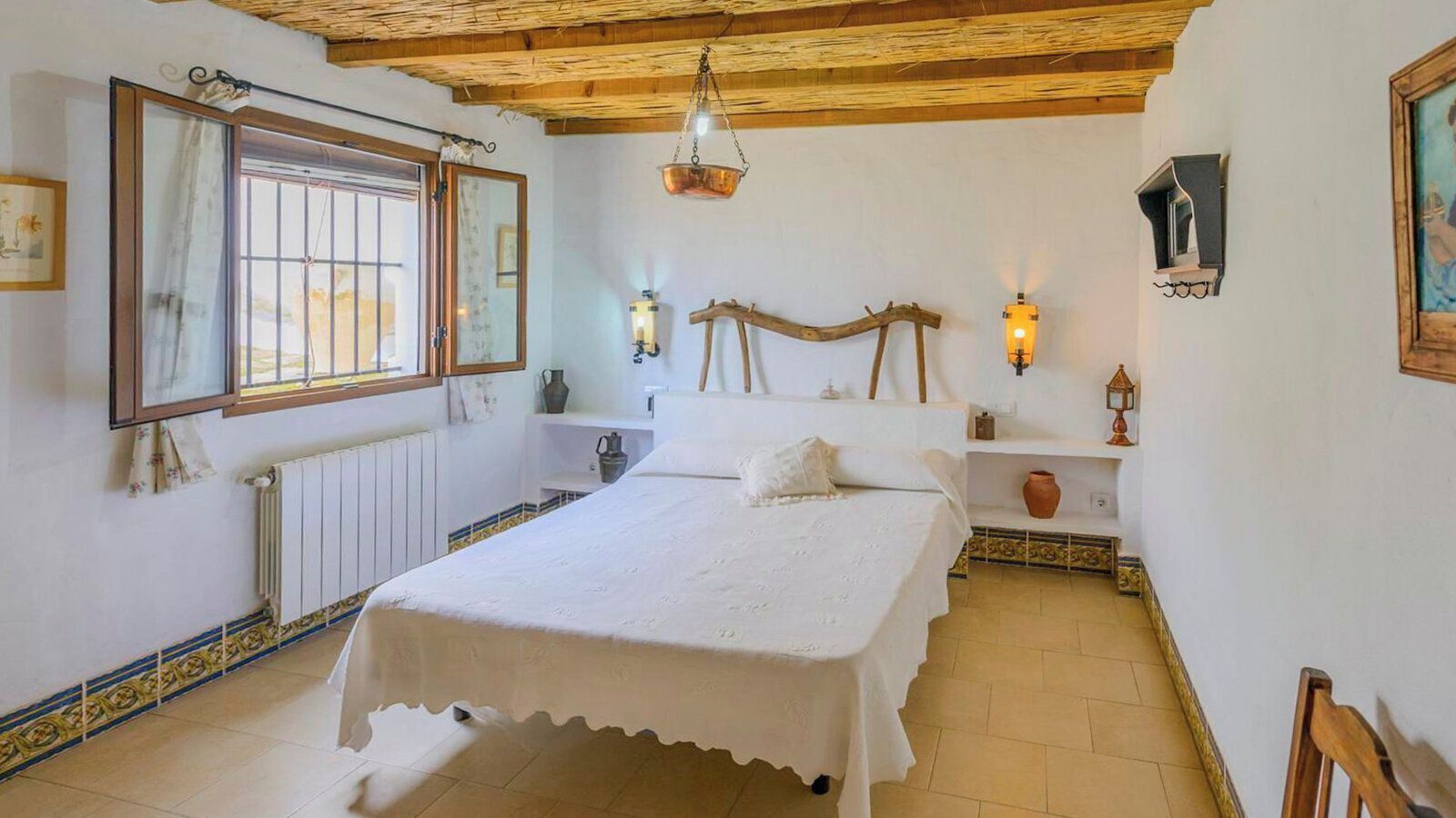 Een slaapkamer met een bed, een radiator, een raam met tralies, een houten plafond en wandlampen.