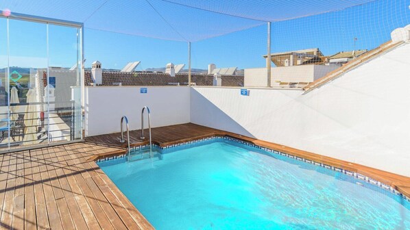 A rooftop swimming pool with clear blue water, wooden deck, and glass walls offering a view of the surrounding buildings.